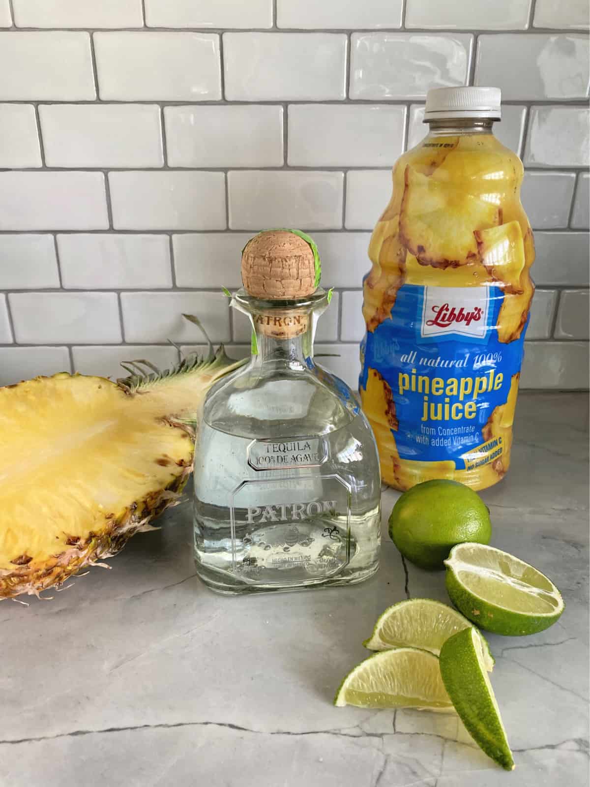 Ingredients on marble countertop; pineapple juice, patron, limes, and fresh pineapple.