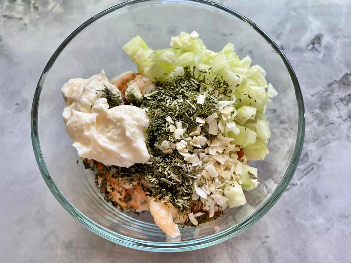 Glass bowl on a marble countertop filled with mayonnaise, shrimp, cucumber, and spices.