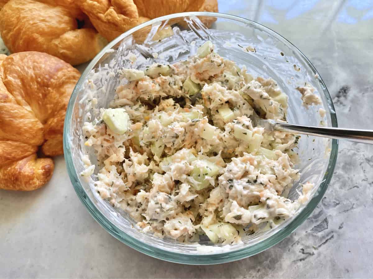 Glass bowl filled with creamy salad resting on a marble countertop with croissants.