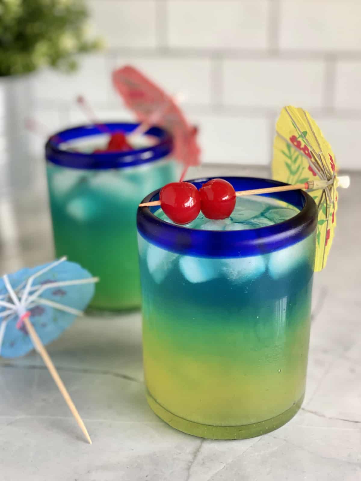 Two drinks that are yellow, green, blue with 2 maraschino cherries on top with a paper umbrella.