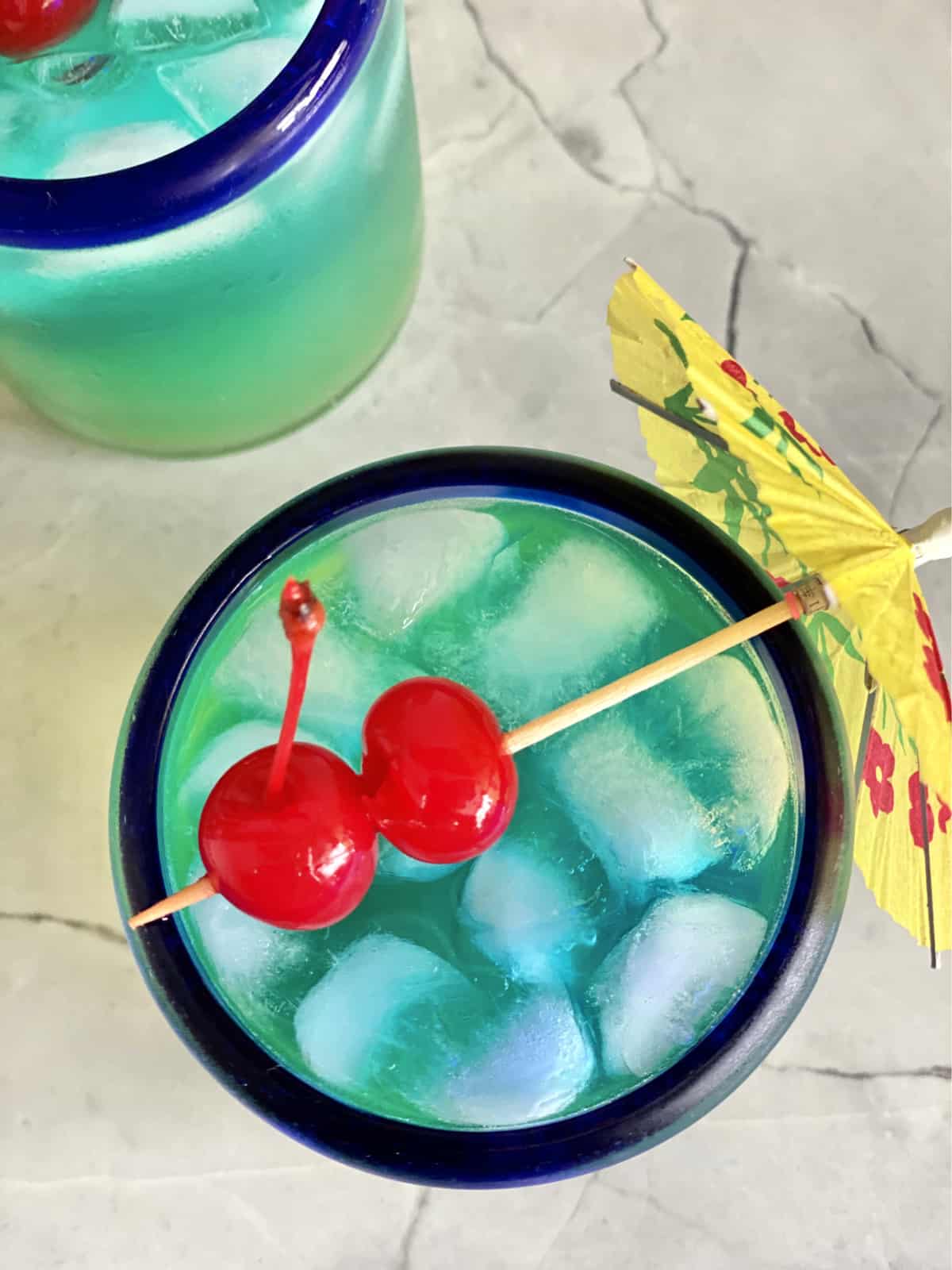 Top view of a blue drink with 2 maraschino cherries on top strung through a paper umbrella.