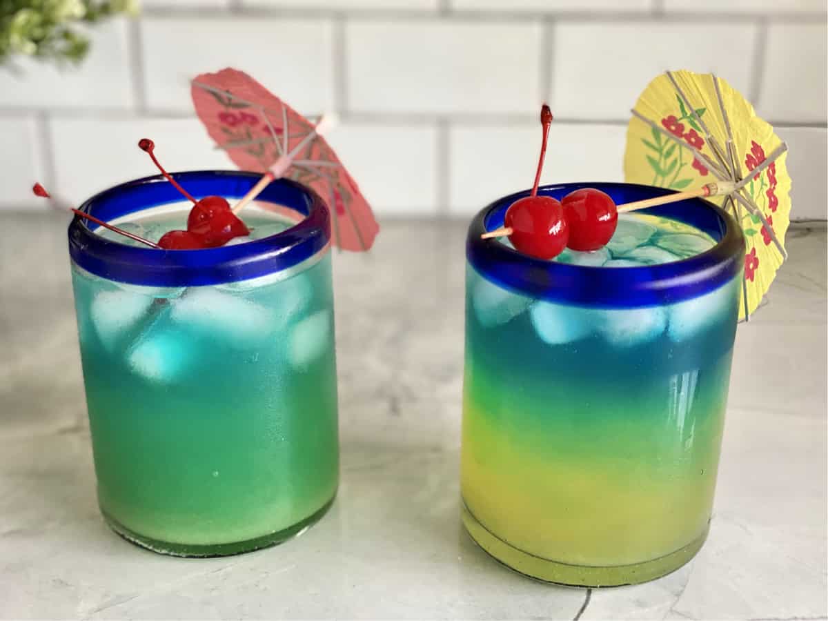 Two drinks side by side one blended light blue color the other layered yellow, green, blue. 