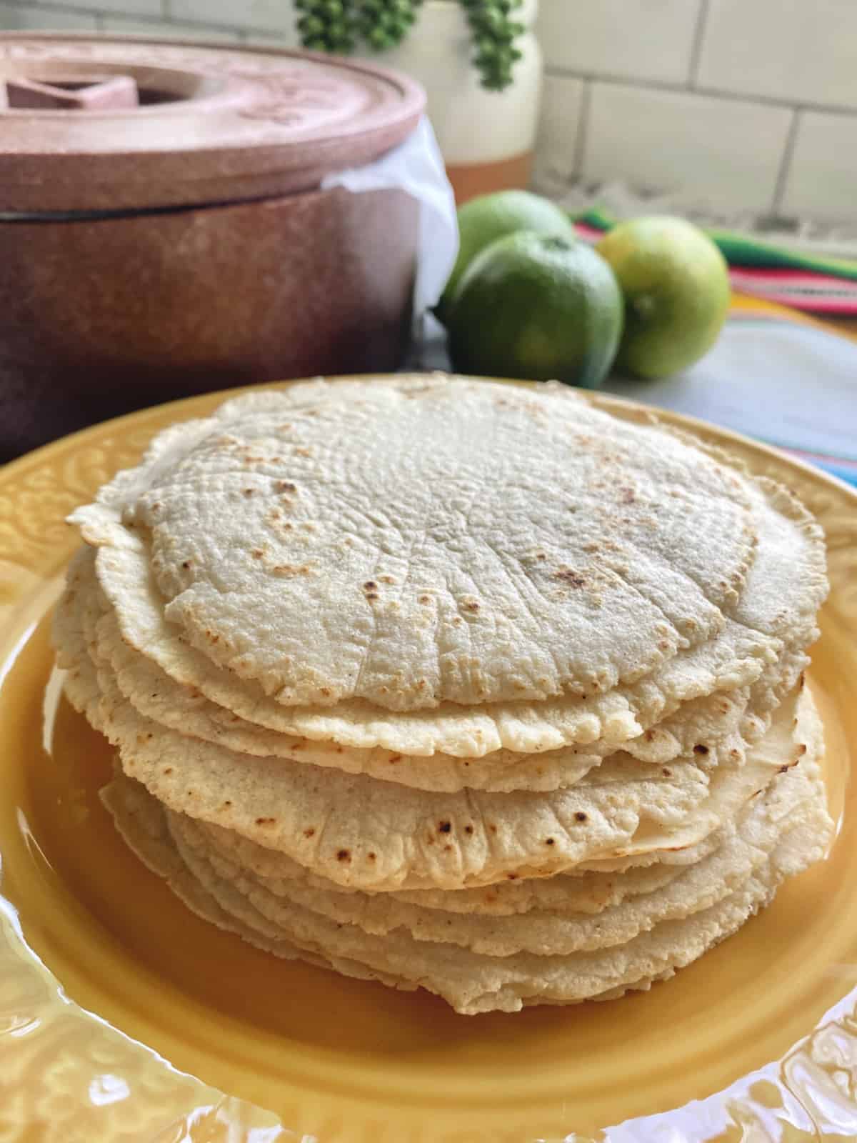 Stack of corn tortillas on a yellow plate with a brown tortilla warmer in the background.