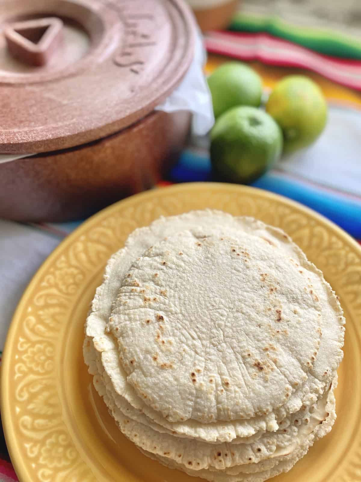 Stack of tortillas on a yellow plate with tortilla warmer in background. 