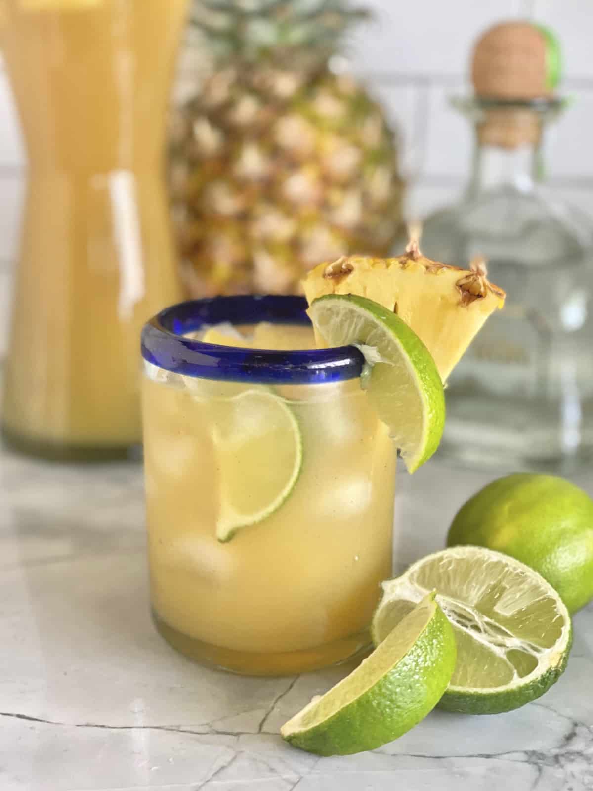 Glass with yellow drink with blue rim garnished with lime and pineapple chunk.