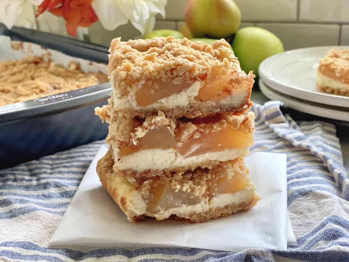 Side view of three apple cheescake bars stacked on top of each other with a pan in the background.