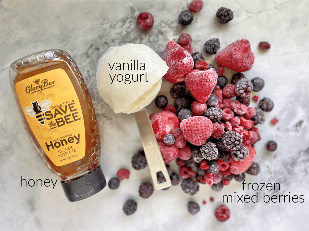 Ingredients on marble countertop: frozen mixed berries, vanilla yogurt, and honey. 