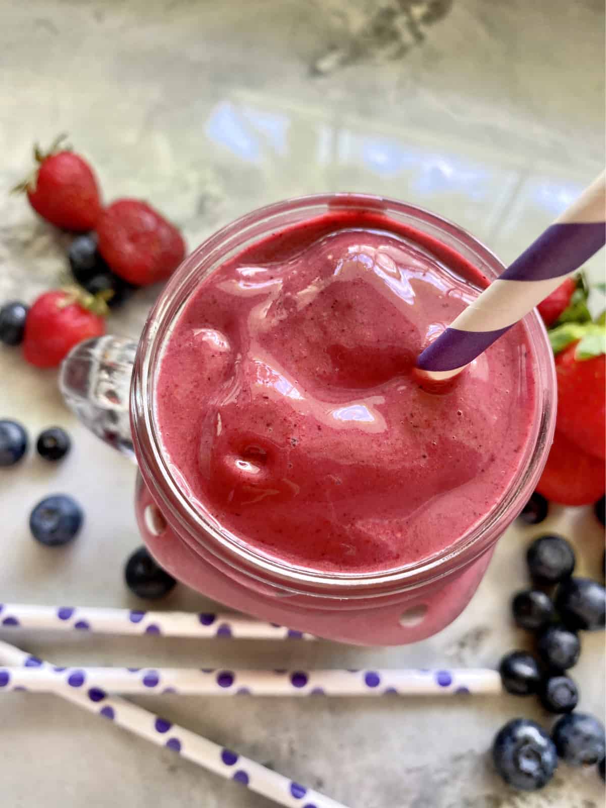 Top view of a purple smoothie in a glass with purple and white stripped straw on marble countertop.