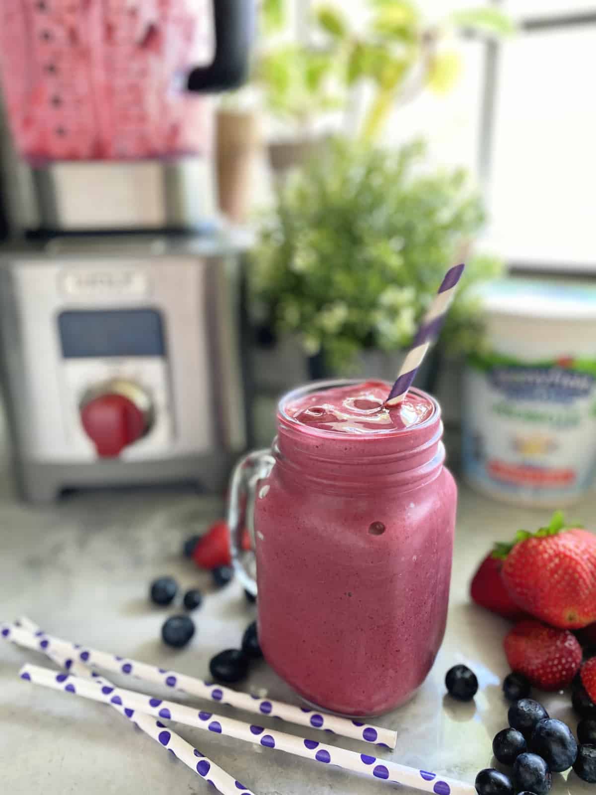 Mason jar glass filled with a purple smoothie with berries and straws on marble countertop. 