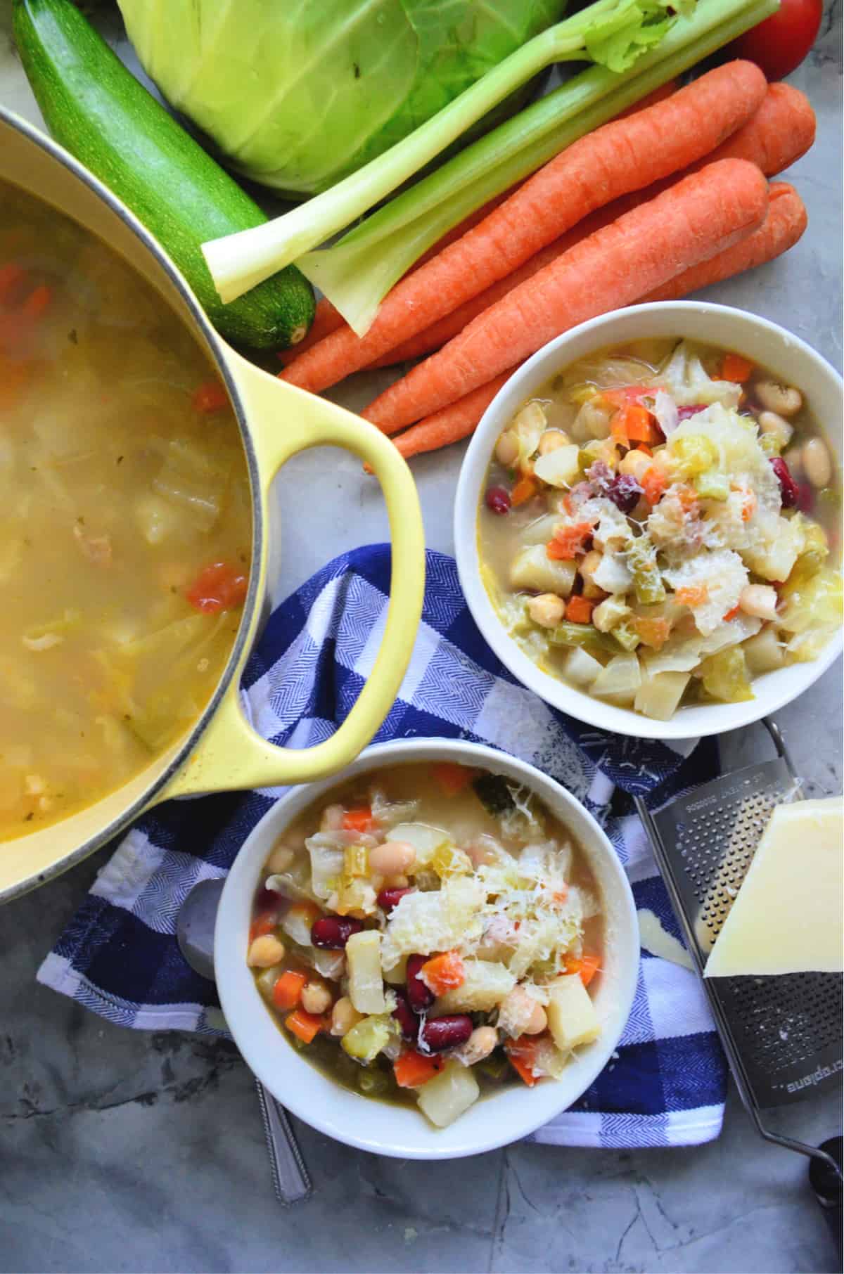 Top view of two bowl so minestrone soup, yellow pot filled with soup, and vegetables on countertop.