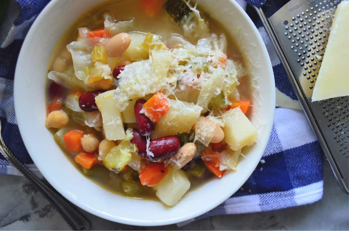 Top view of a white bowl filled with minestrone soup with cheese grater and cheese next to it.