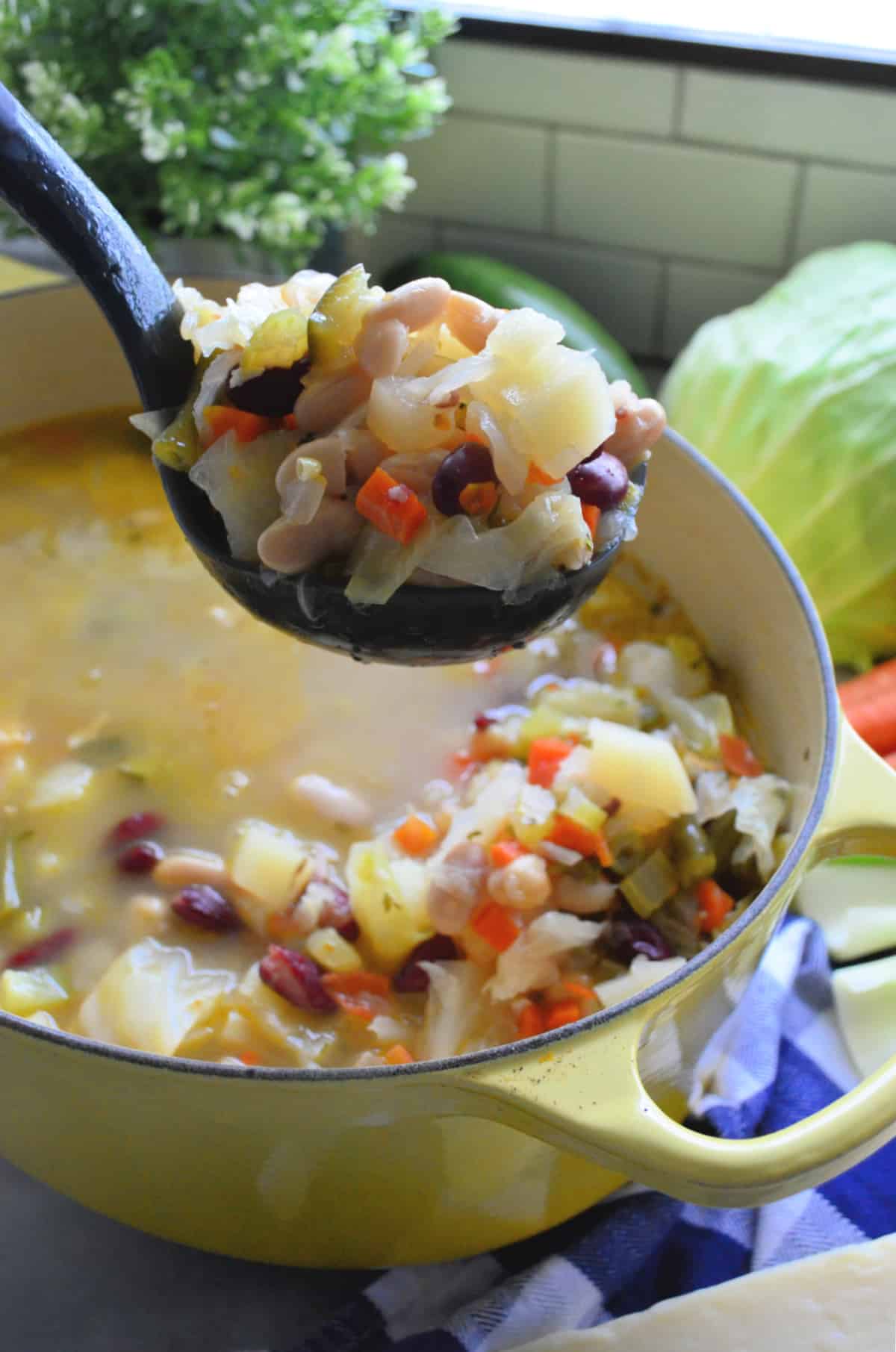 Close up of a black ladle filled with minestrone soup over a yellow pot.