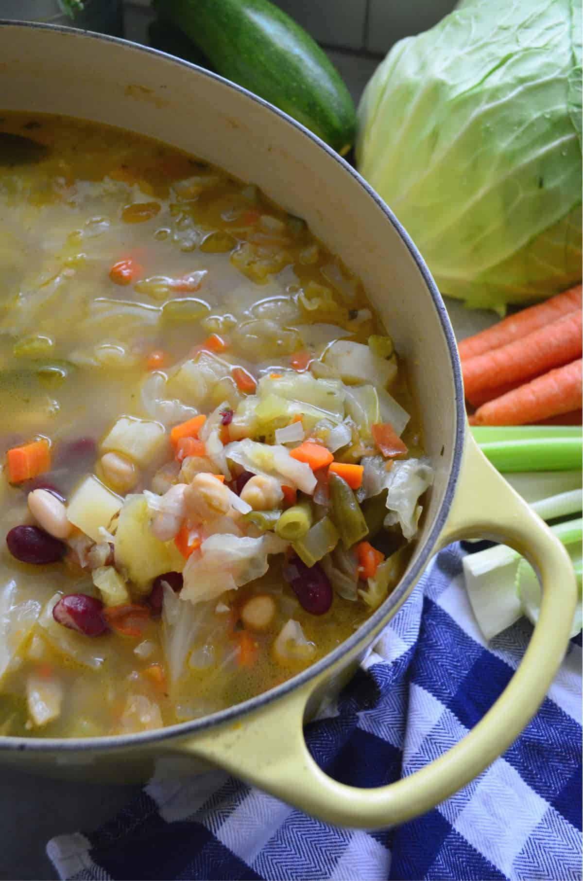 Top view of yellow pot filled with minestrone soup with vegetables in background.