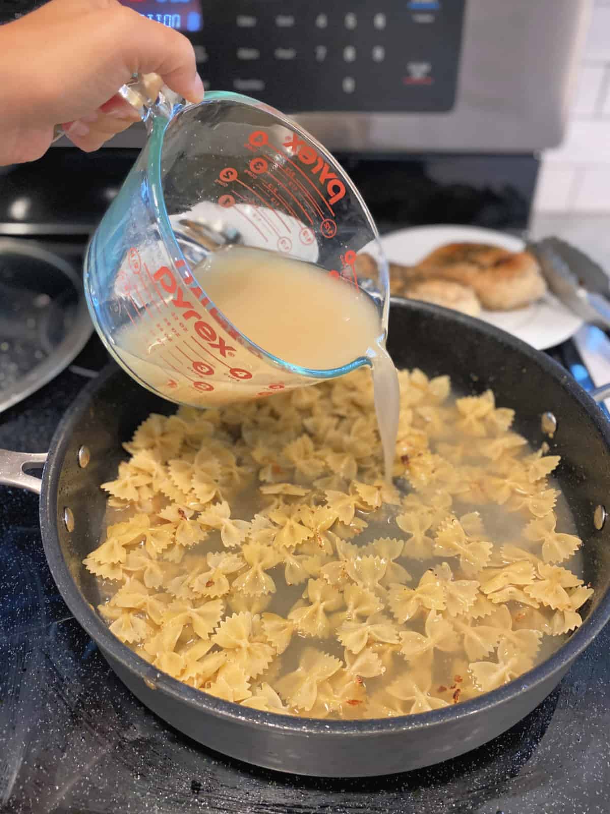 Female hand pouring chicken stock into a black skillet filled with bow tie pasta.