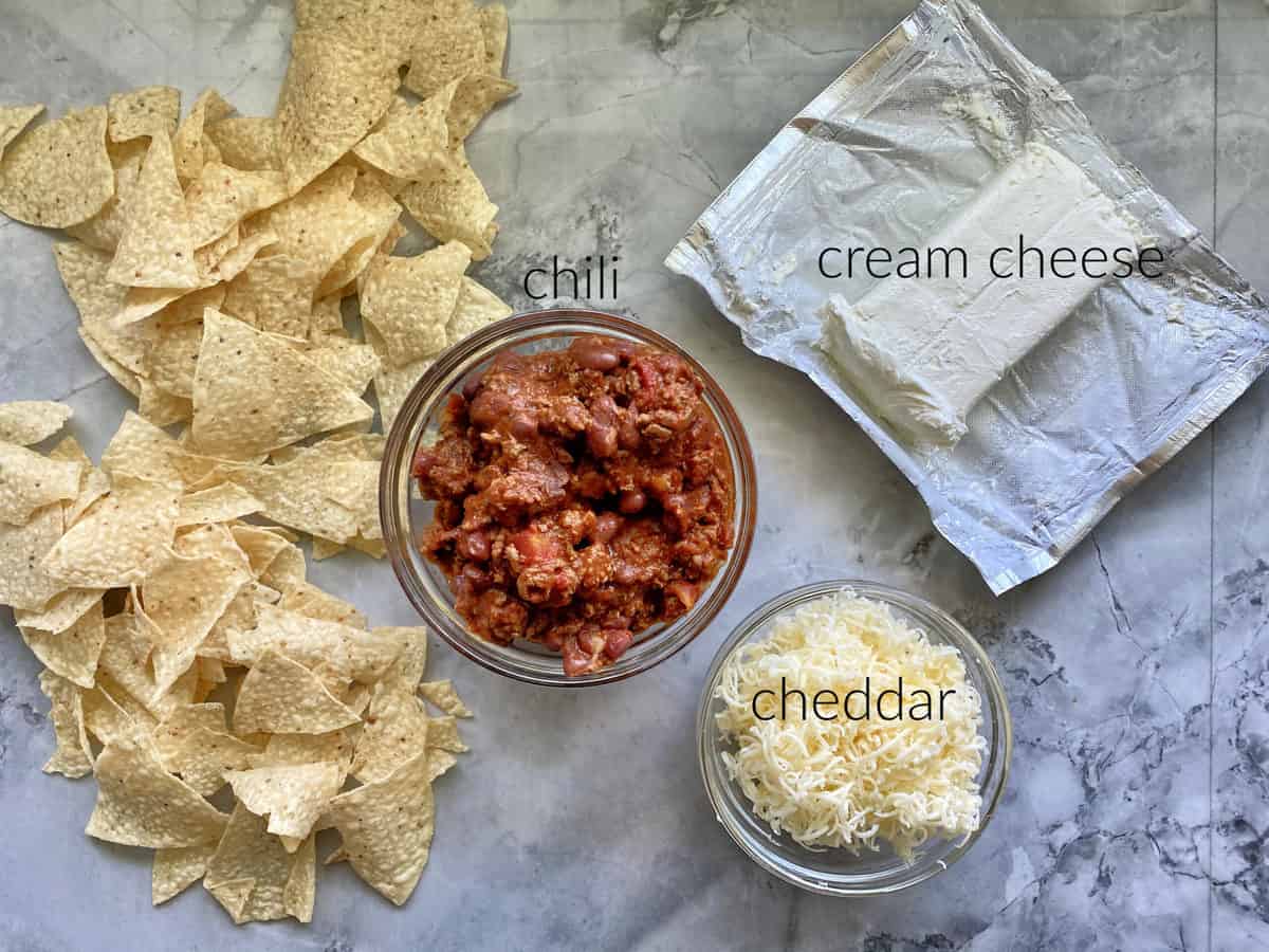 Ingredients on marble countertop: tortilla chips, chili, cream cheese, cheddar cheese.