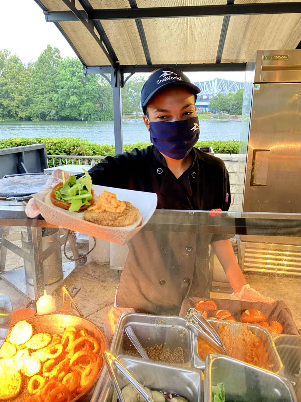 SeaWorld Orlando employee wearing mask, hat, and gloves handing a fried green tomato slider over counter.