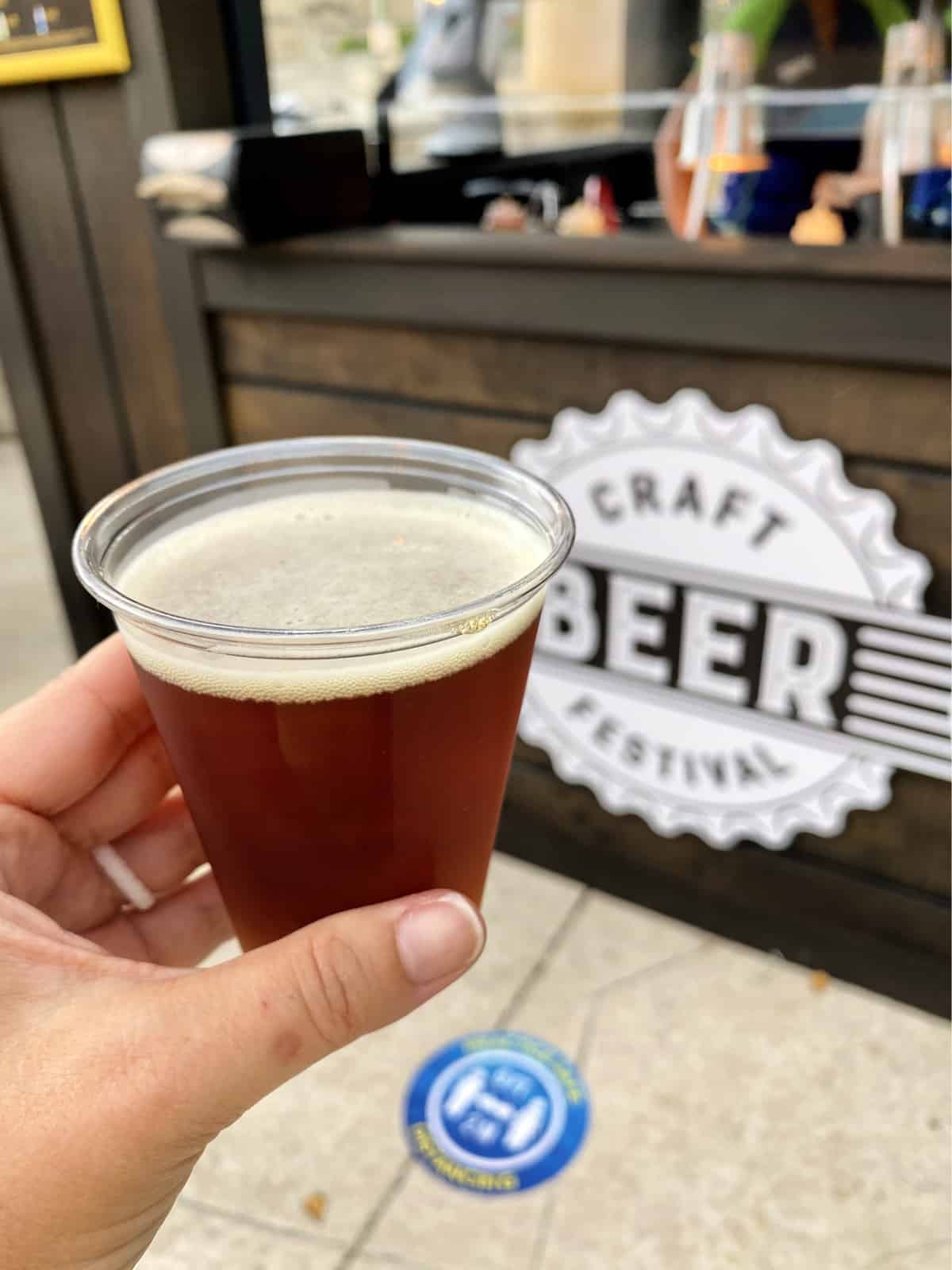 Female hand holding a small cup of brown ale beer with a craft beer festival sign in the background.