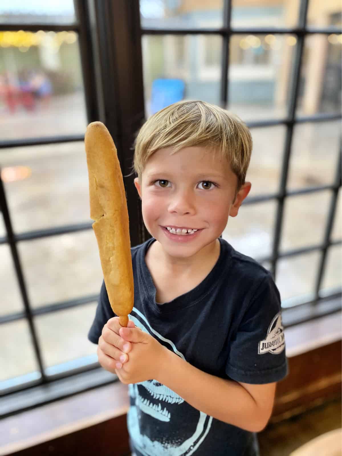 Little blonde haired boy in black shirt holding a foot long corn dog.