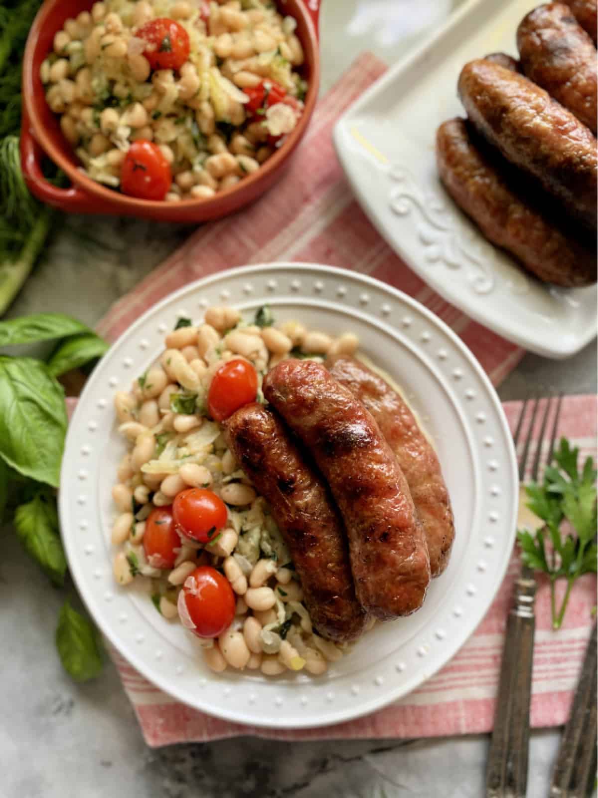 Top view of a white plate with white beans and sausage, platter of sausage and white beans on the side.