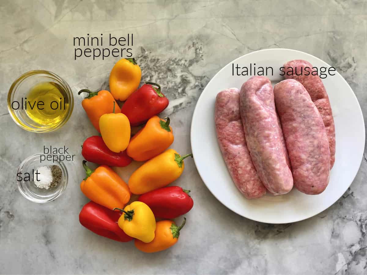 Ingredients on a marble countertop: olive oil, salt, pepper, mini bell peppers, Italian sausage.