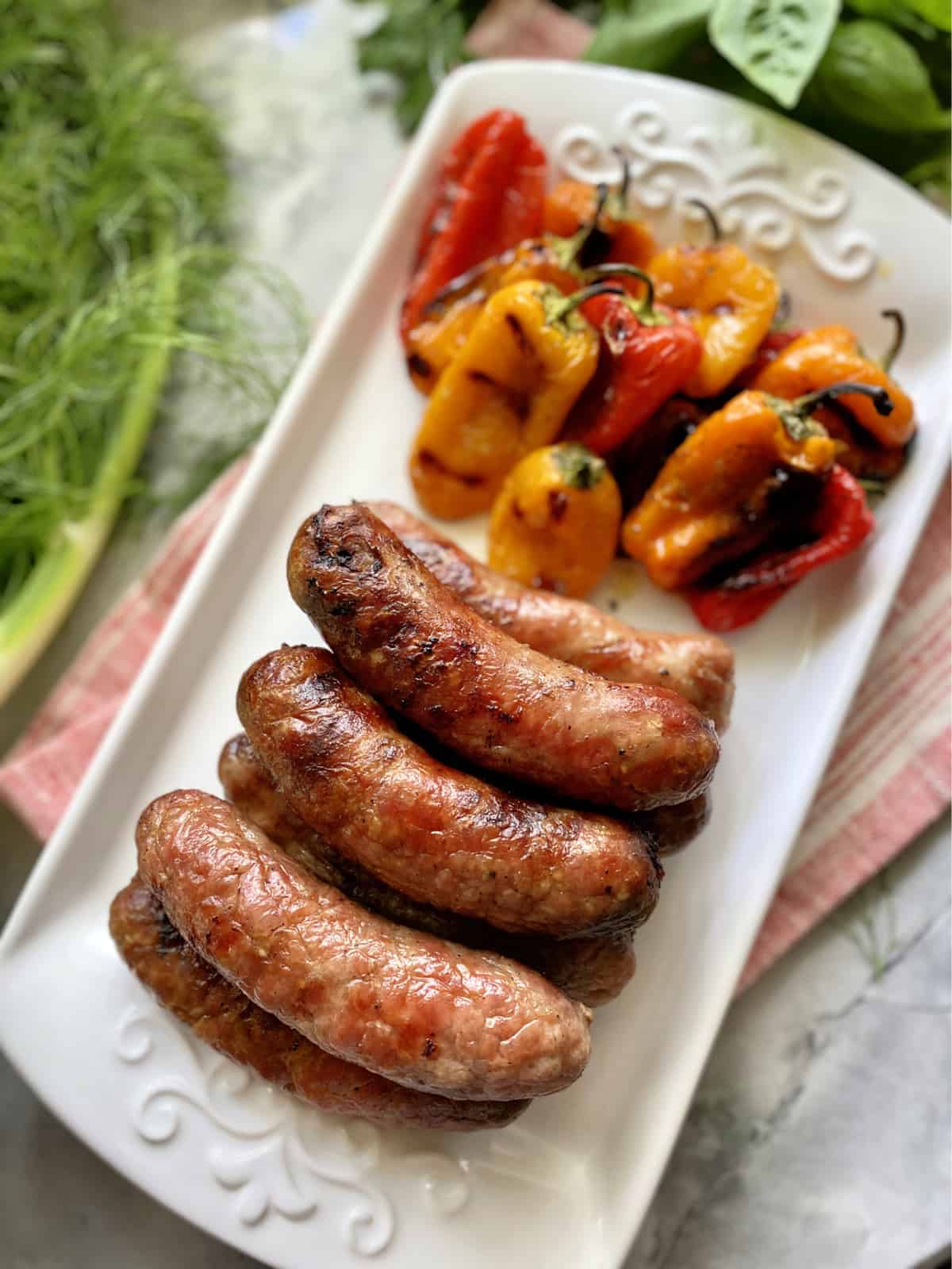 Top view of grilled Italian sausage with charred mini peppers on a white plate with fresh herbs.