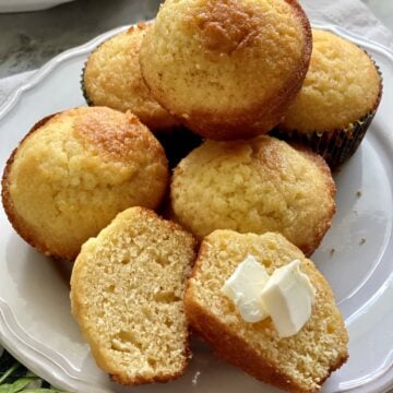 White plate with 5 muffins stacked and one corn muffin split with butter on top.