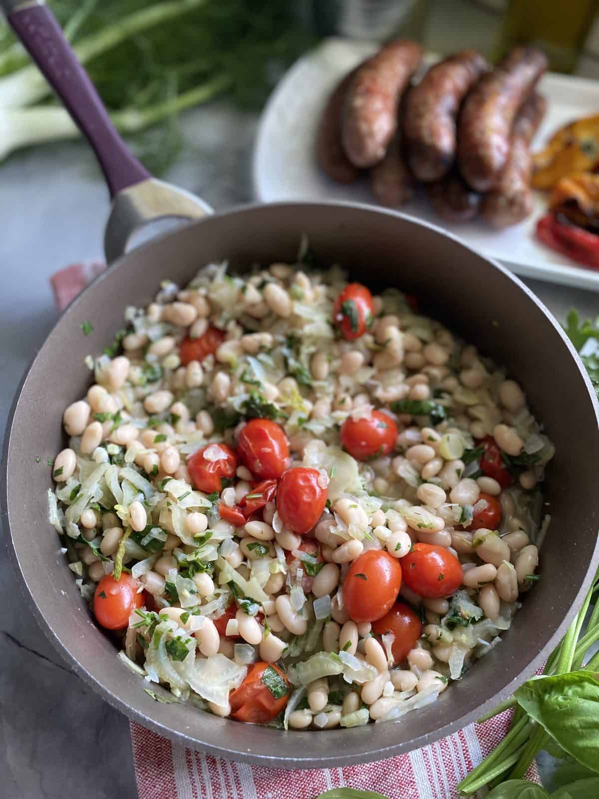 Brown skillet filled with white beans, fennel, and tomatoes with fresh herbs.
