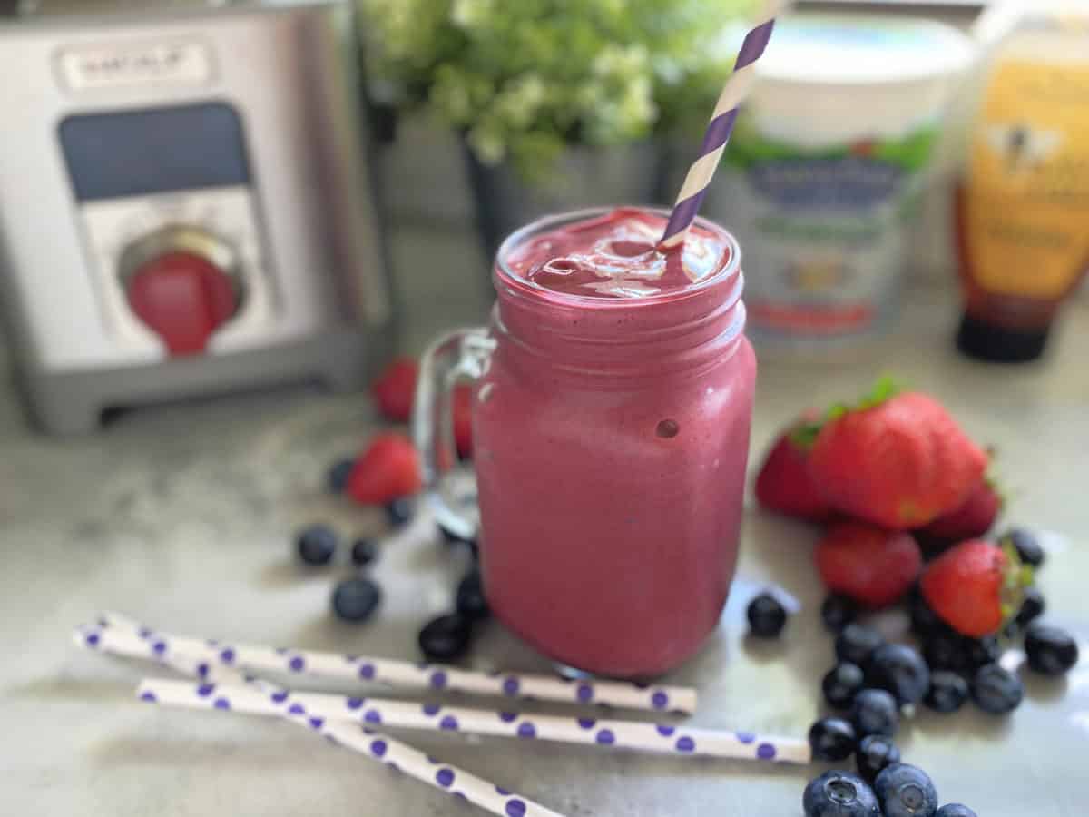Purple smoothie in a glass mug with striped purple and white straw with berries on counter.