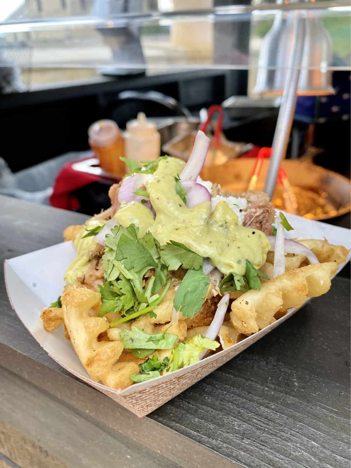 Waffles fries in a paper tray topped with pork, cilantro, red onions, and green cream sauce.