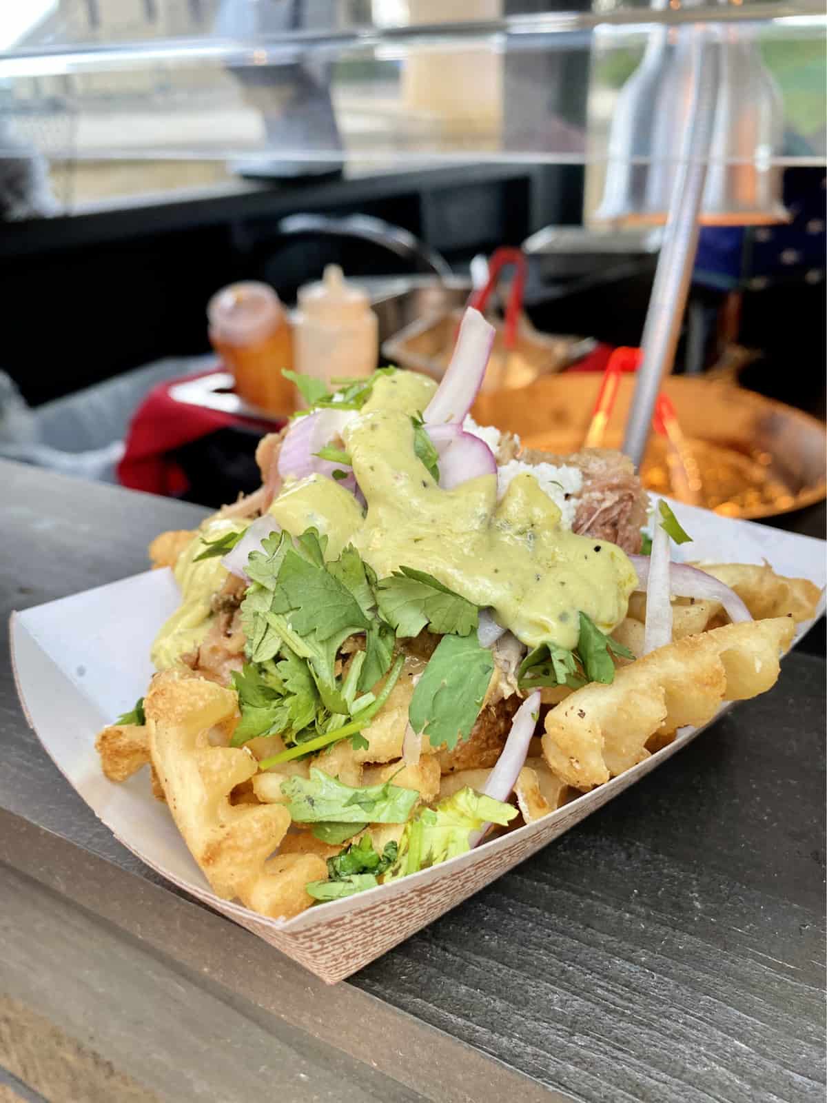 Waffles fries in a paper tray topped with pork, cilantro, red onions, and green cream sauce.