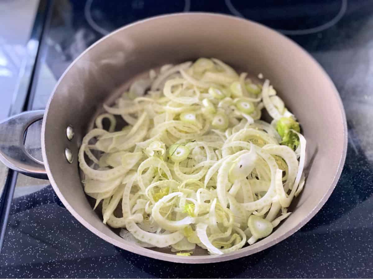 Brown saute pan with shaved fennel and steam rising from pan on stove.