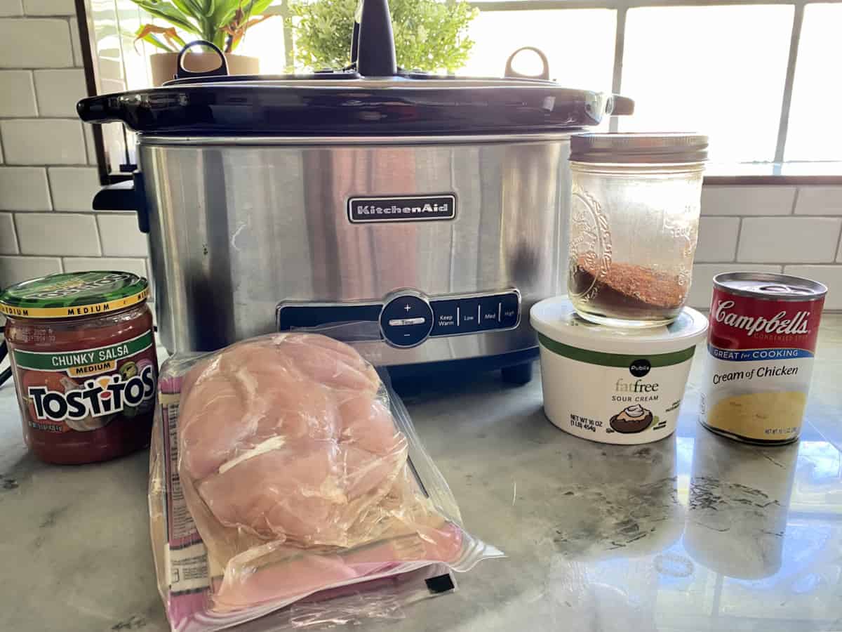 Ingredients on countertop with slow cooker: chicken breast, taco seasoning, cream of chicken soup, sour cream, salsa