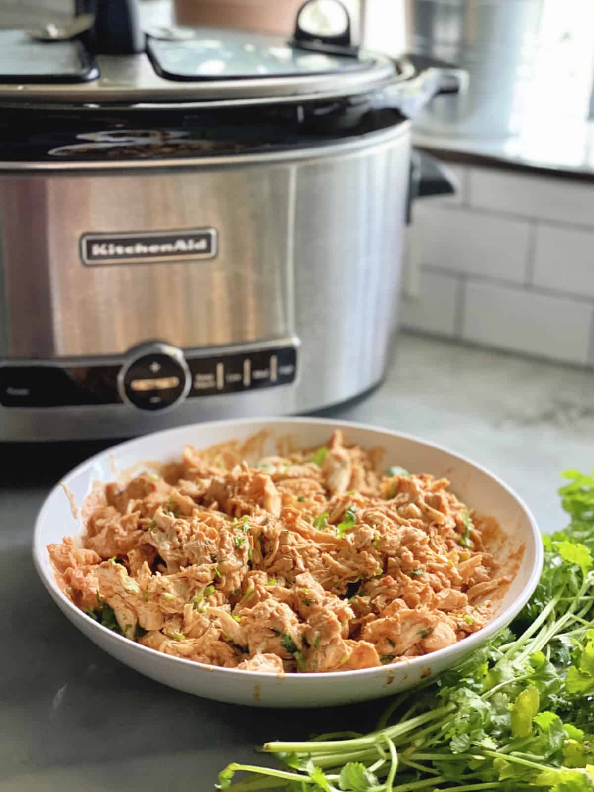 Shredded Chicken with cilantro in a white bowl with slow cooker in background.