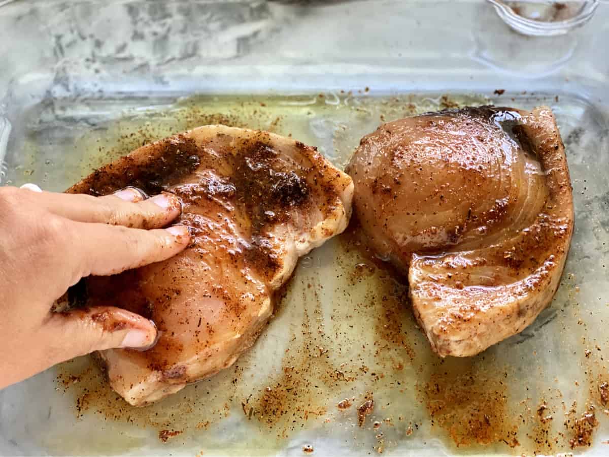 Two swordfish steaks on a glass dish with blackening seasoning and olive  slathered on it.