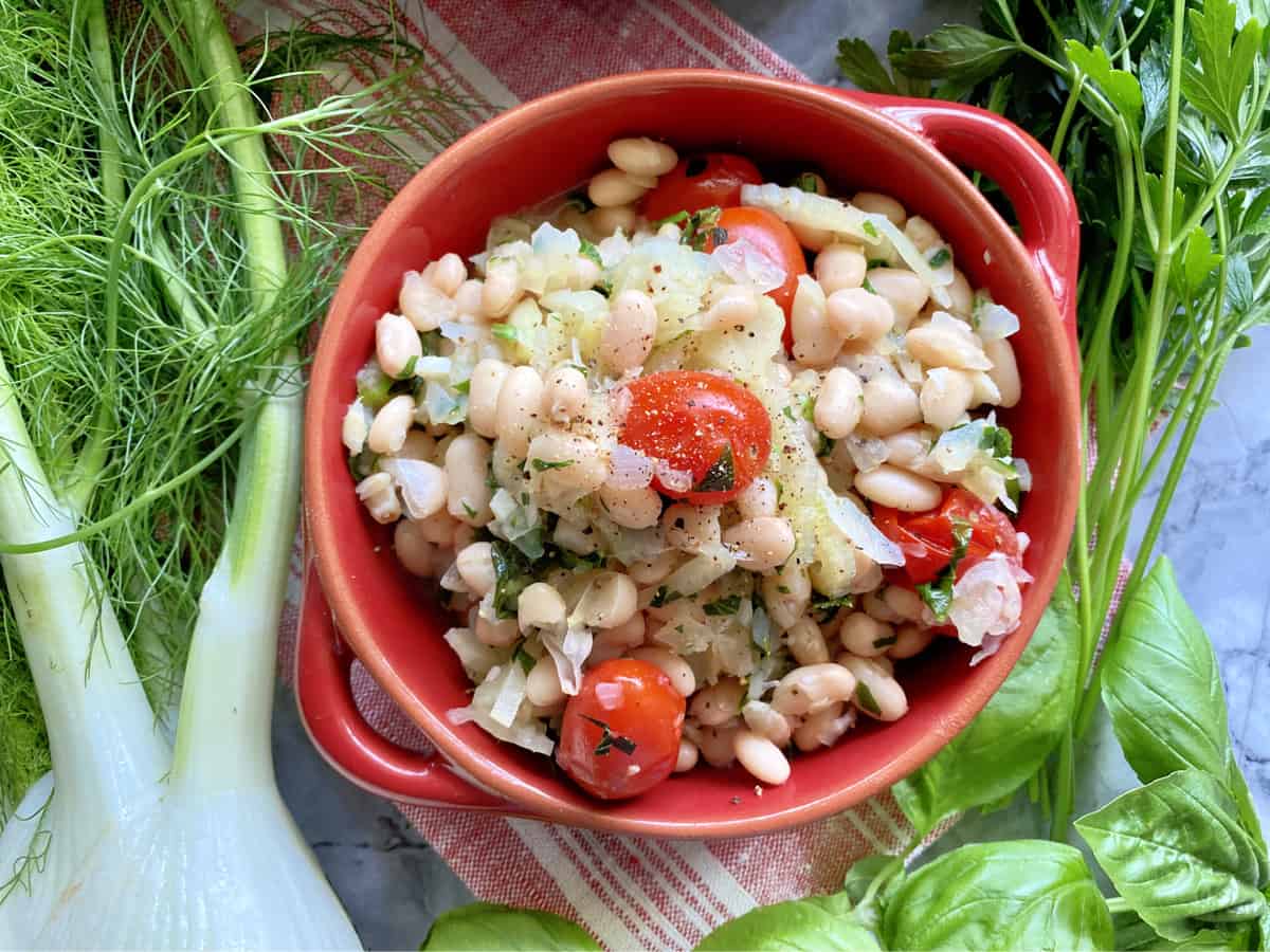 Top view of a red round dish holding white beans with grape tomatoes and fennel on the side.