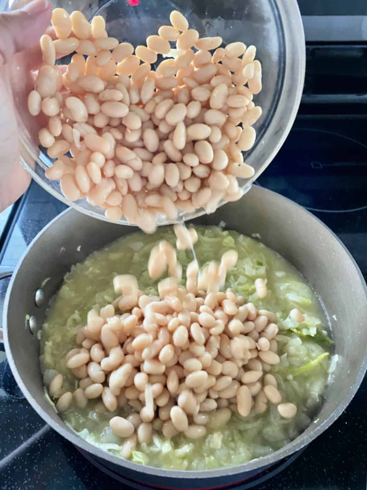 Female hand holding glass bowl, pouring white beans into skillet.