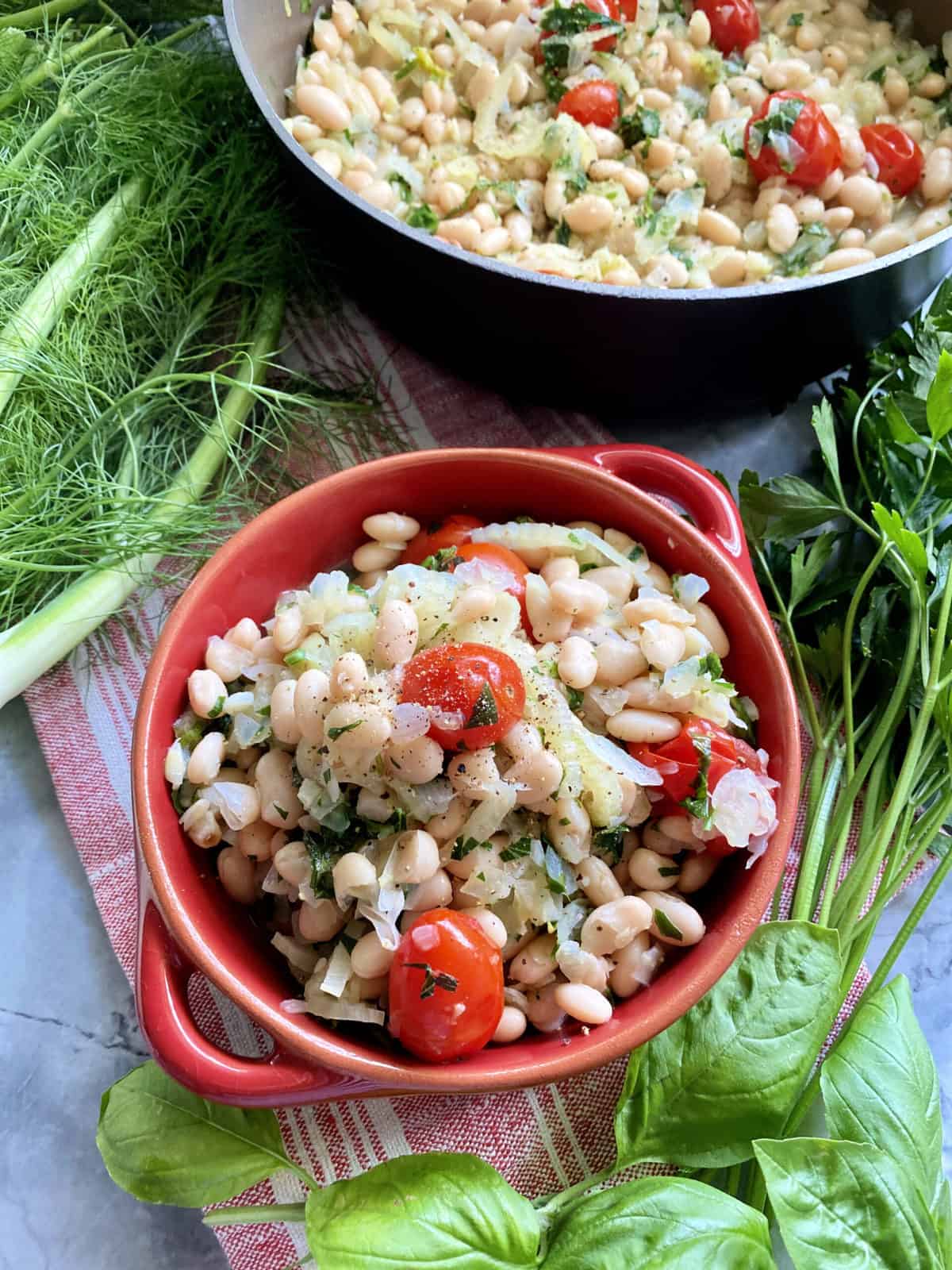 Top view of a red dish with white beans and tomatoes and skillet full of white beans.