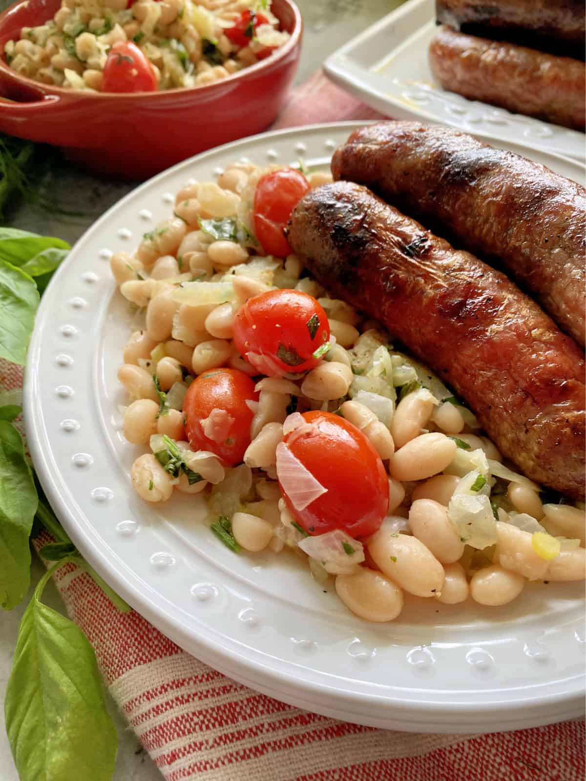White plate with white beans and tomatoes with fresh herbs and grilled sausage links.