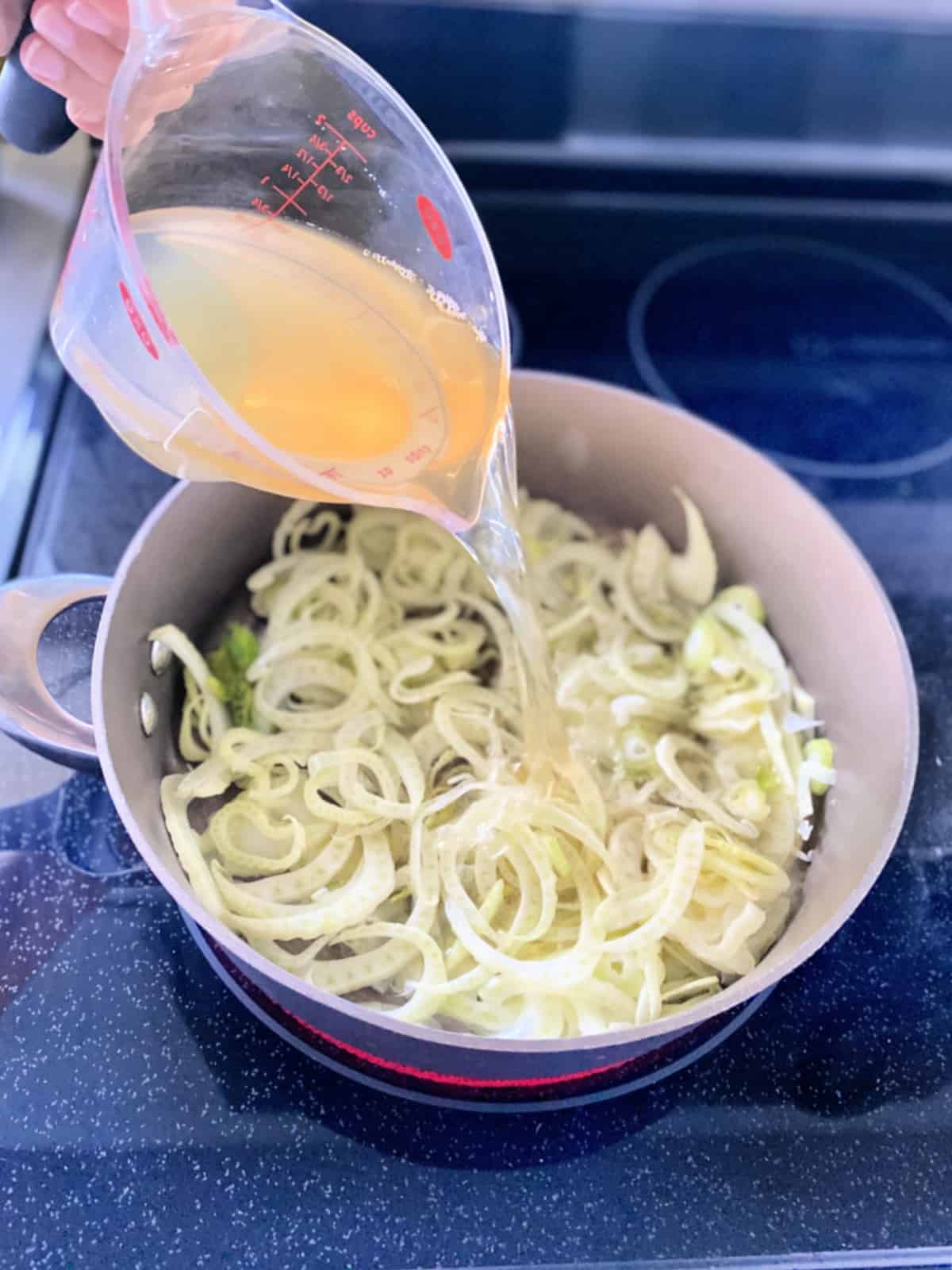 Female hand pouring chicken broth via measuring cup into a skillet with sliced fennel.