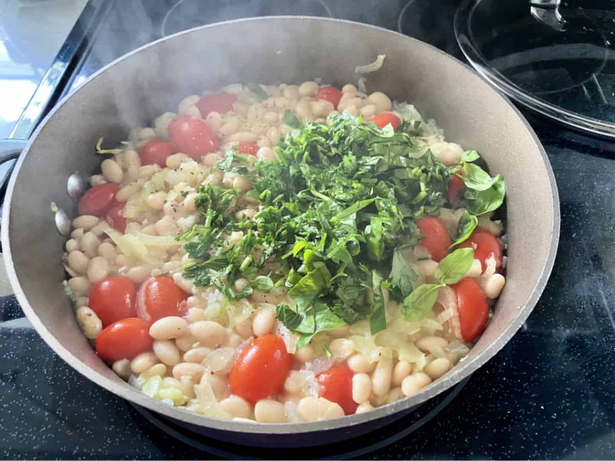 Brown skillet with white beans, grape tomatoes, and fresh herbs cooking with steam rising.