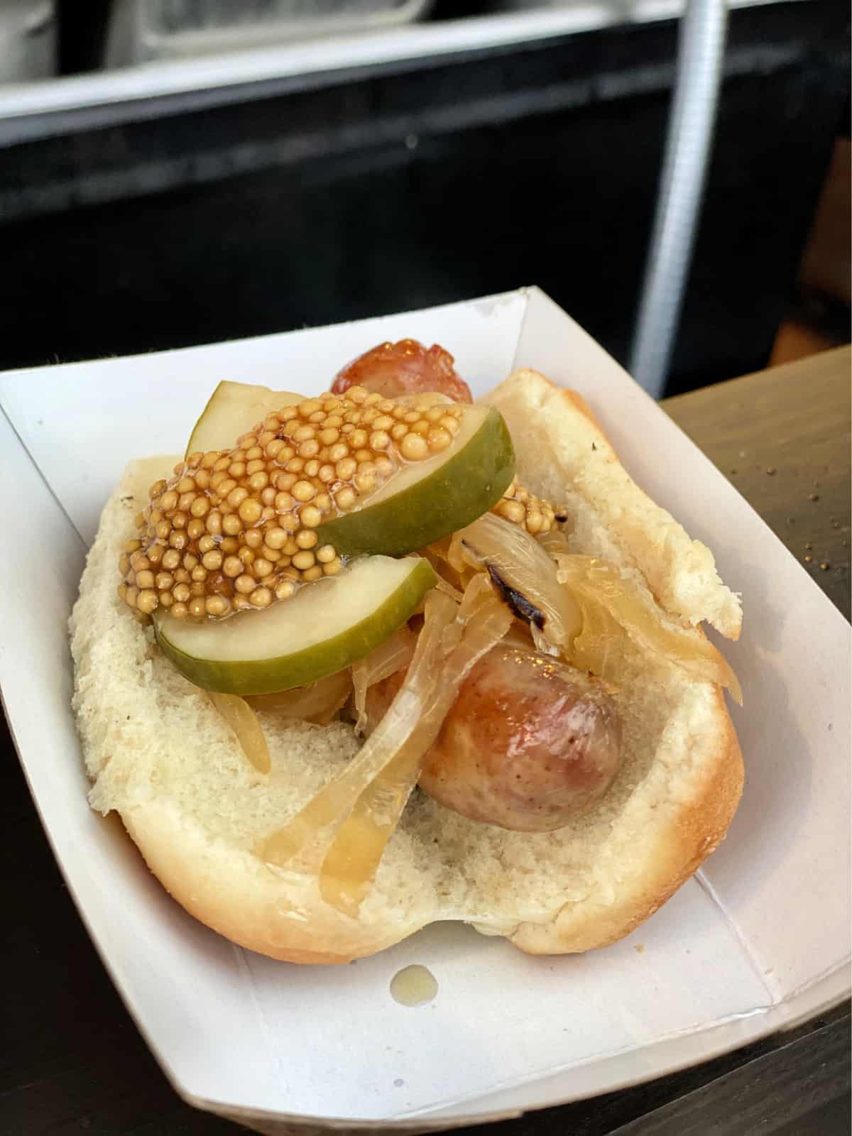 Sausage in a bun with caramelized onions, pickles, and grainy mustard in a paper tray.