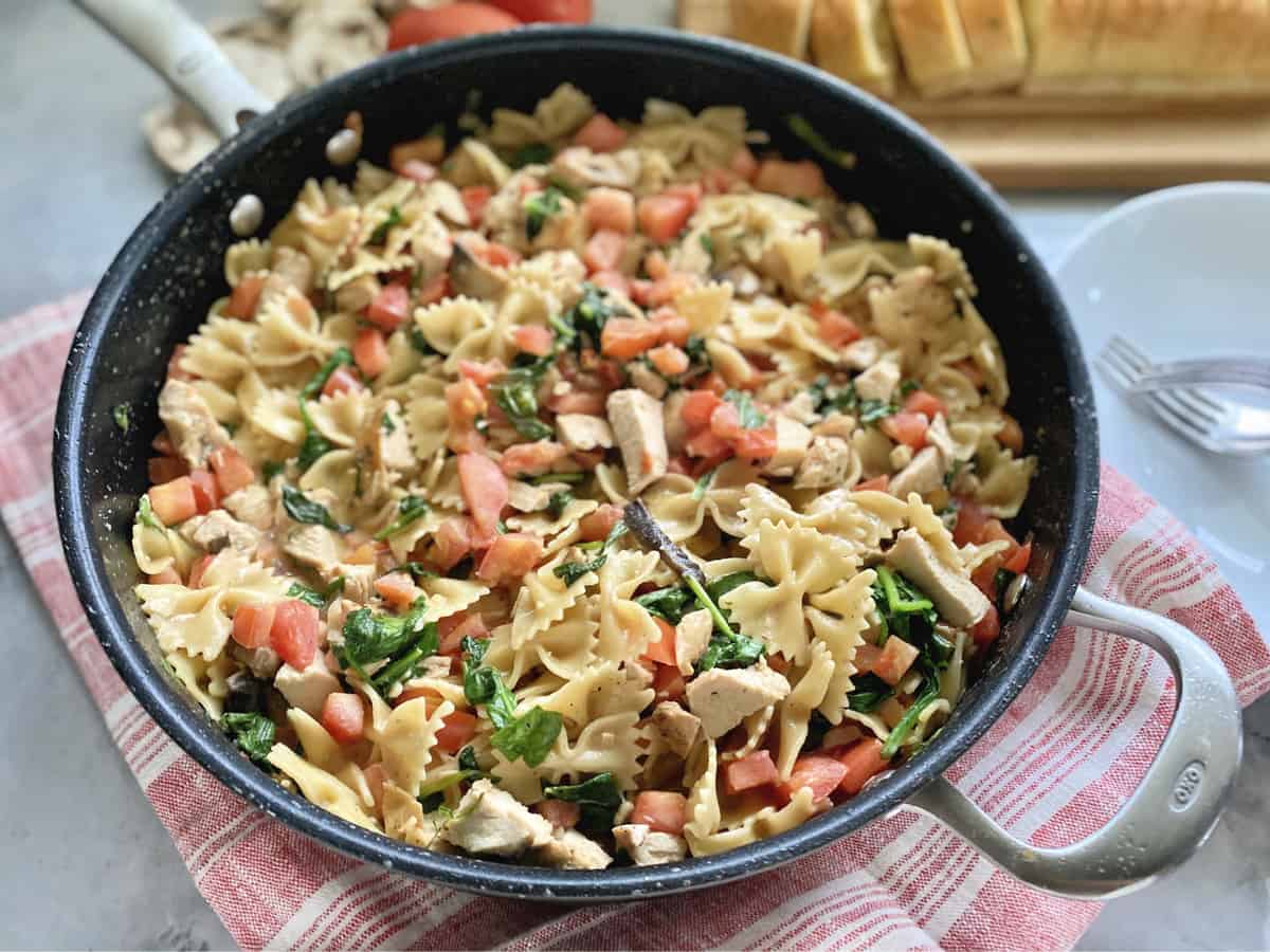 Skillet on red and white towel filled with bow tie pasta, chicken, tomatoes, and spinach.