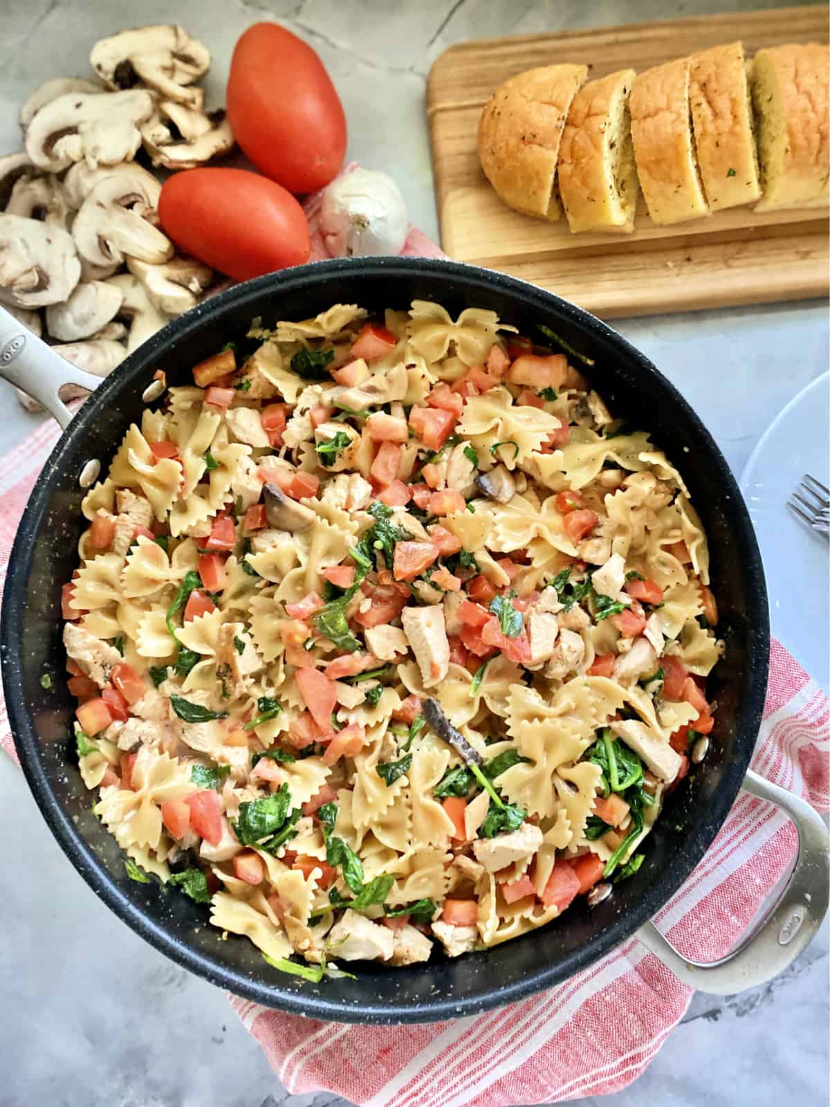 Top view of a large OXO black skillet filled with bow tie pasta, chicken, spinach and tomatoes.