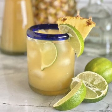 Glass cup with yellow liquid with a lime and pineapple wedge on the glass rim.