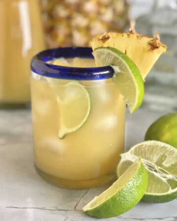 Glass cup with yellow liquid with a lime and pineapple wedge on the glass rim.