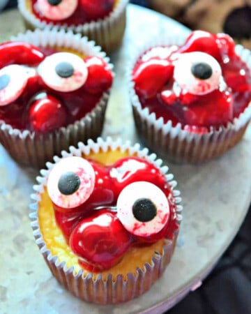 four cherry cheeseacke cupcakeson a metal platter with candy eyeballs on top.