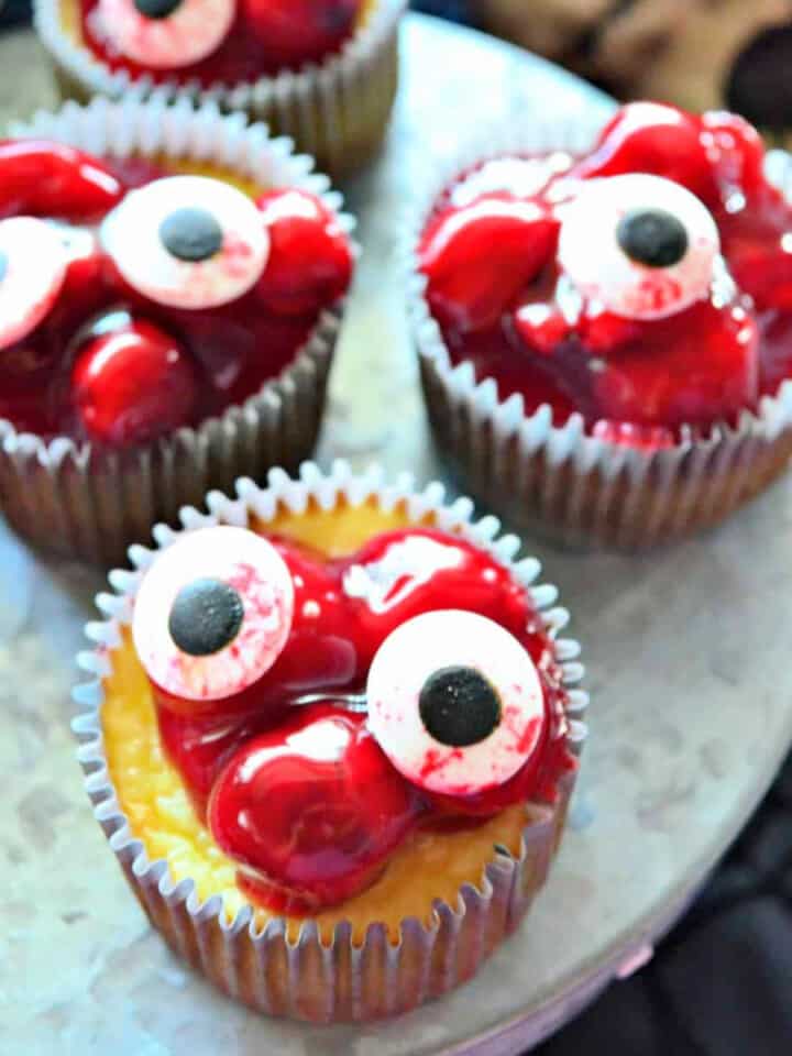 four cherry cheeseacke cupcakeson a metal platter with candy eyeballs on top.