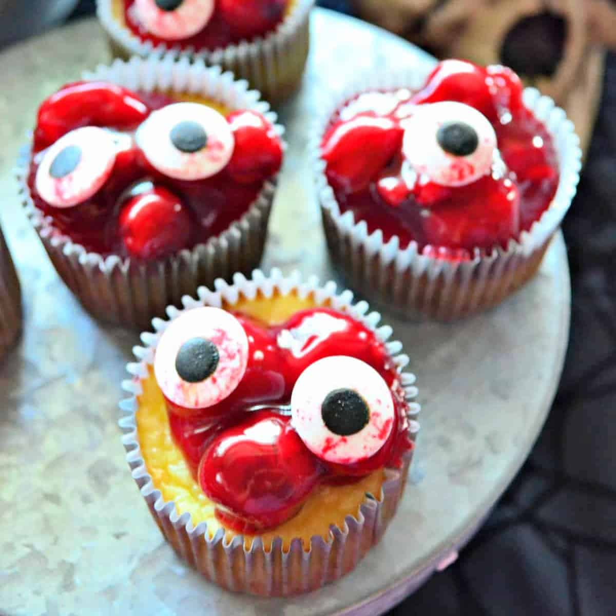 four cherry cheeseacke cupcakeson a metal platter with candy eyeballs on top.