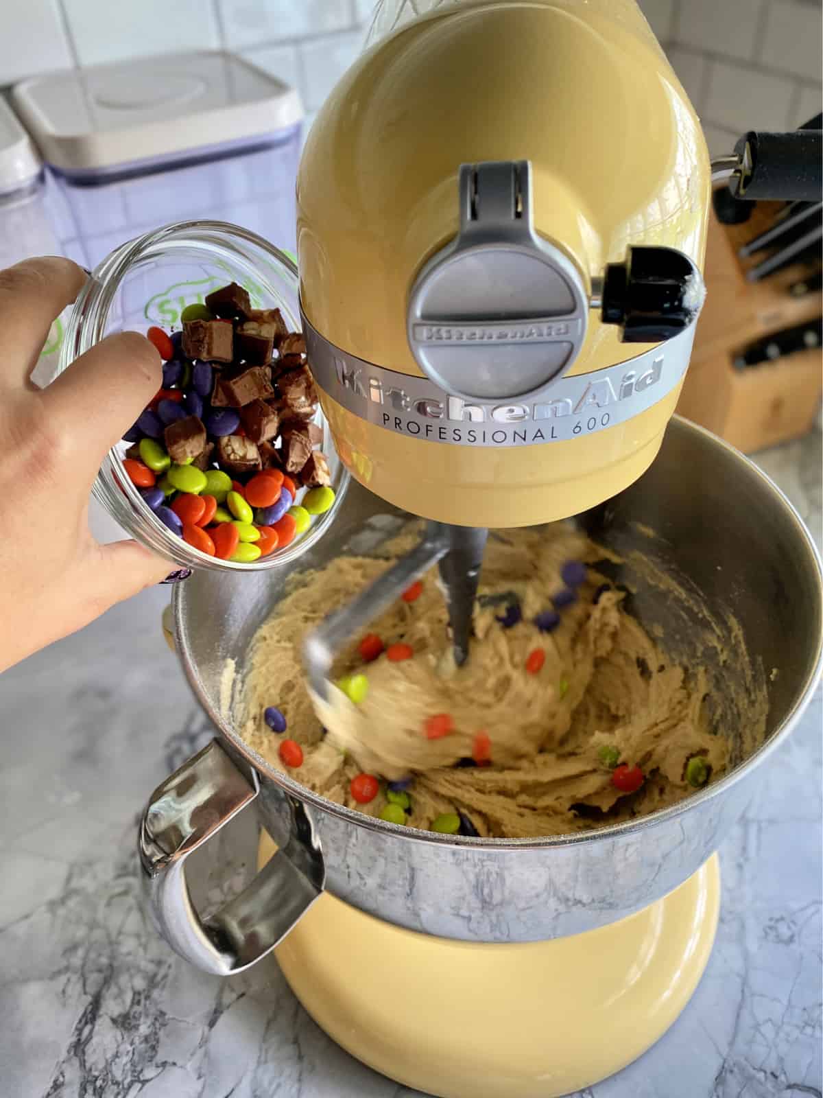 Candy being poured into a yellow KitchenAid Mixing bowl with cookie dough.