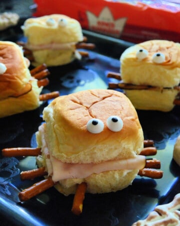 Four mini sandwiches that look like spiders with candy eyes and pretzels sticking out the sides.